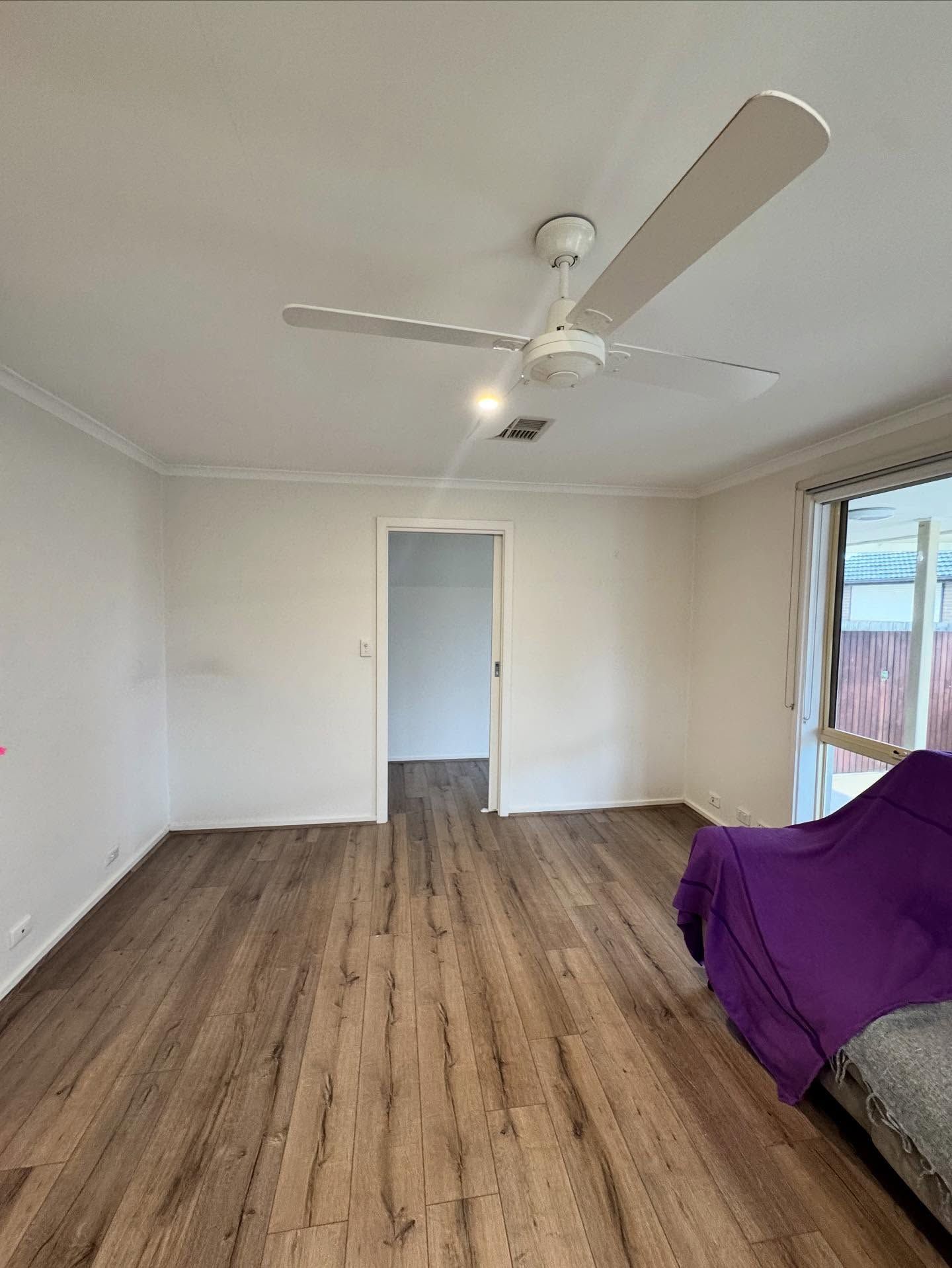 Empty room with wood floor, white walls, ceiling fan, doorway, and window; purple-covered couch at right.