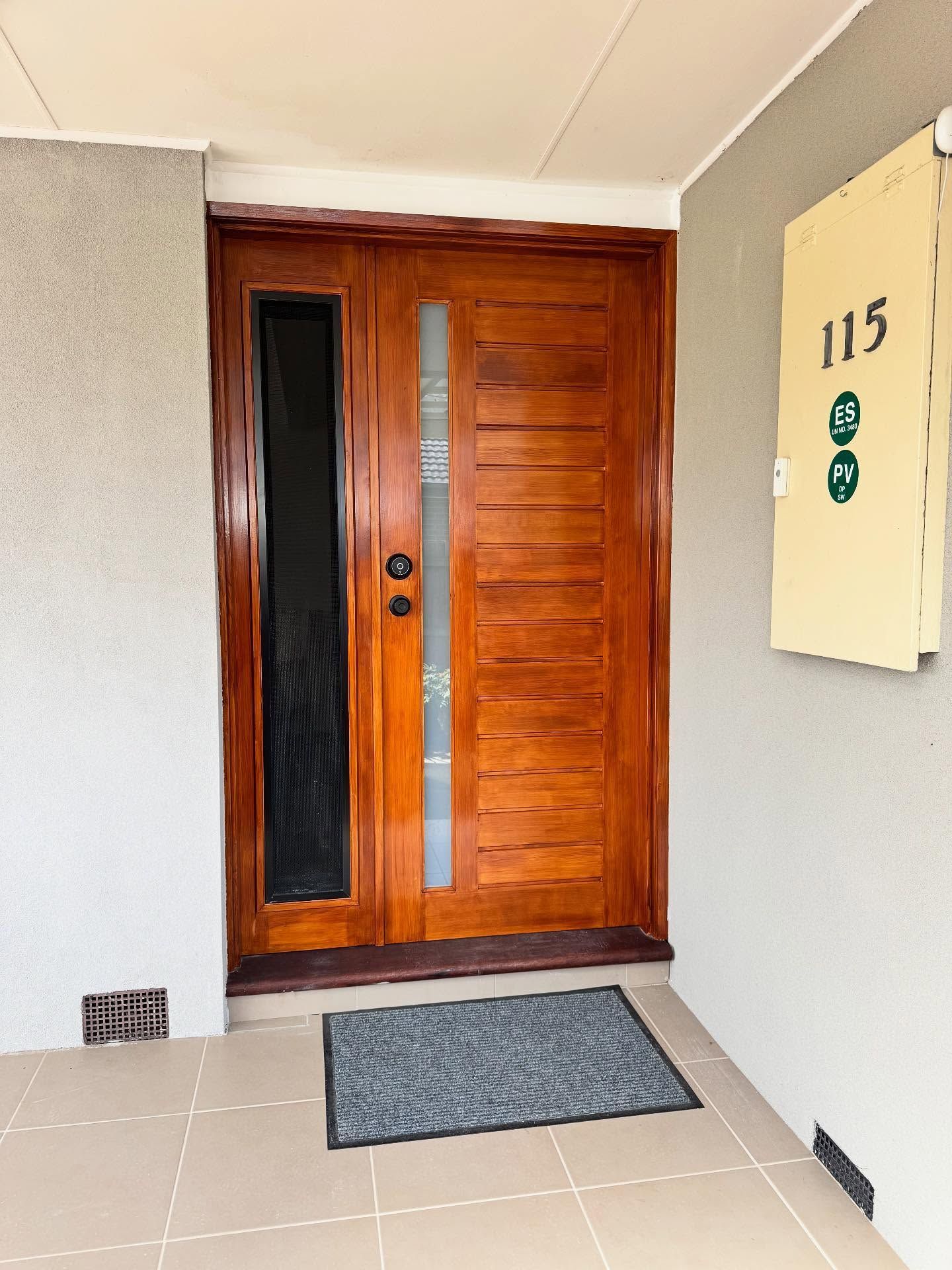 Wooden front door with glass panels and a gray doormat at apartment 115 entrance.