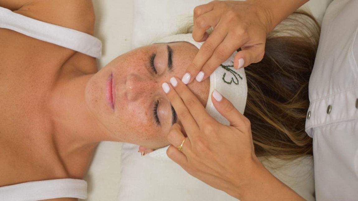 Mujer recibiendo un masaje facial con los ojos cerrados, con una diadema blanca en un entorno de spa.