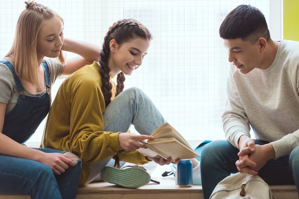 Tre persone guardano un libro vicino a una finestra; una tocca la spalla dell'altra, tutte sorridenti.