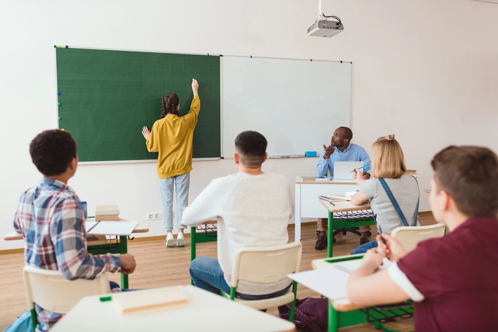 Uno studente scrive su una lavagna in classe; l'insegnante e gli studenti osservano.