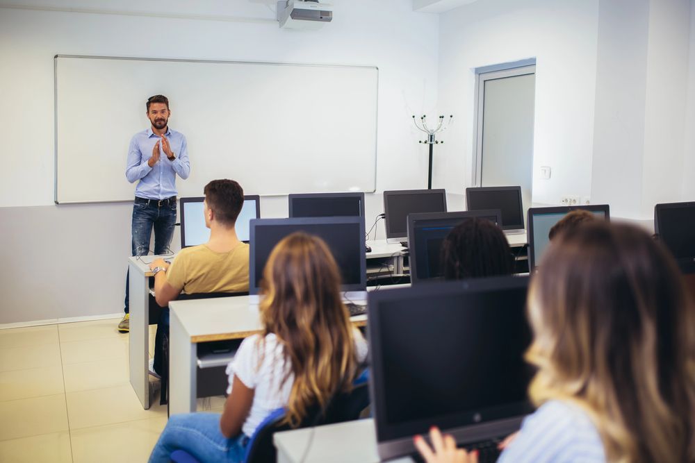 Un uomo tiene una lezione in un laboratorio informatico, gli studenti sono seduti alle scrivanie con i monitor.