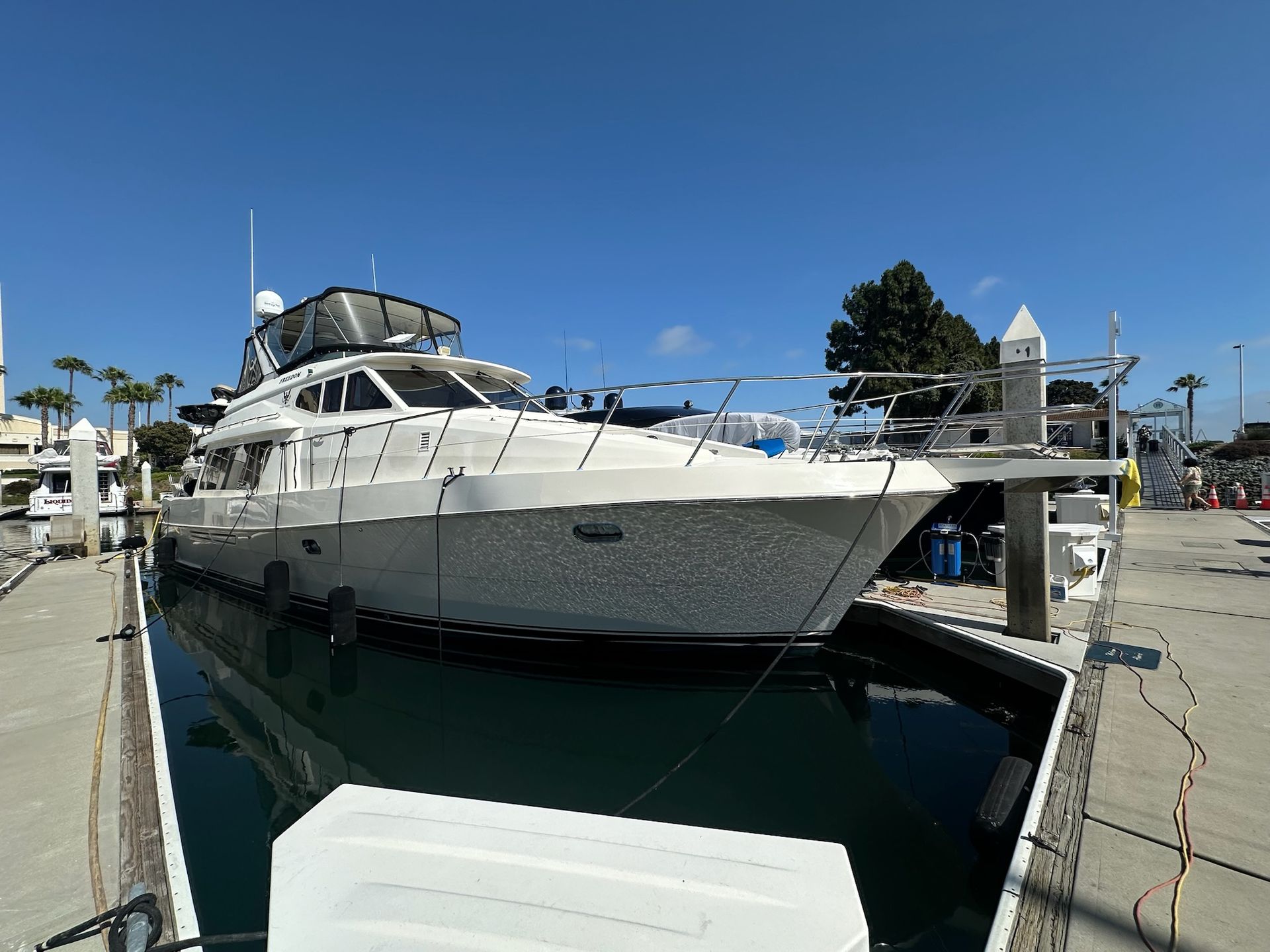 A large white yacht docked at a marina under a bright blue sky. Palm trees and other boats are in the background.
