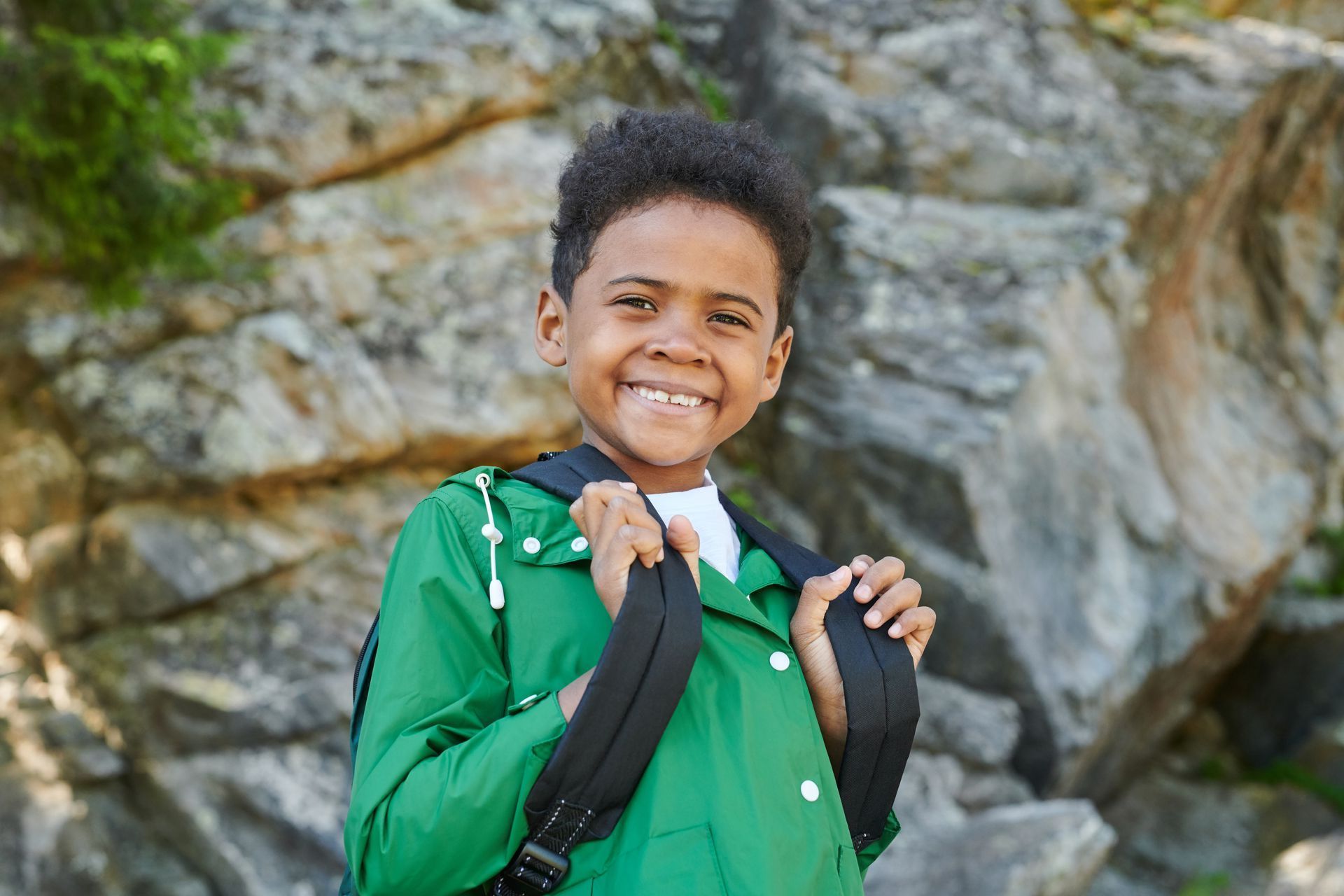 Young boy in front of a rock wearing a green shirt and backpack. He is smiling.