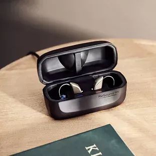 Two ReSound hearing aids in a black charging case on a wooden table, with a book partially visible.