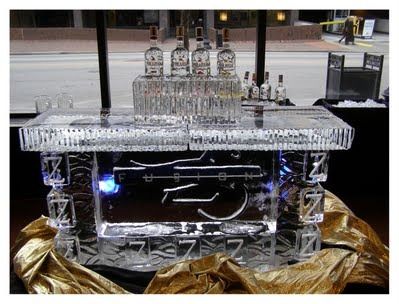 Clear ice sculpture with embedded car design and small bottles on top, displayed on a gold-draped table.