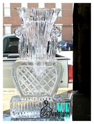 Tall cut-glass vase sculpture with reflective facets displayed in a window against a brick building background