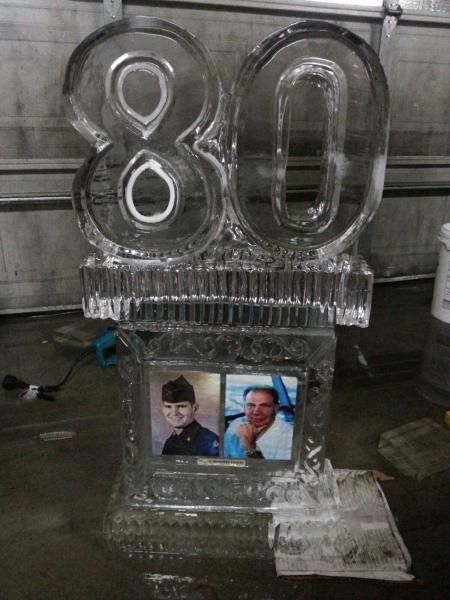 Clear ice sculpture of “80” on a base with two framed photos inside, displayed outdoors.
