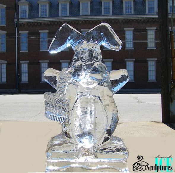 Clear ice sculpture of a rabbit standing outdoors in front of a brick building.