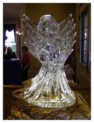 Transparent glass angel sculpture on a pedestal in a museum gallery, lit warmly with gold drapery below