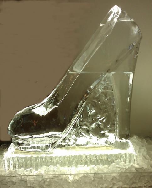 Clear glass high-heel shoe sculpture on a textured base, lit from the side.