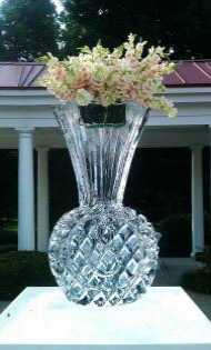 Clear cut-glass vase with pale pink flowers on a white pedestal outdoors.