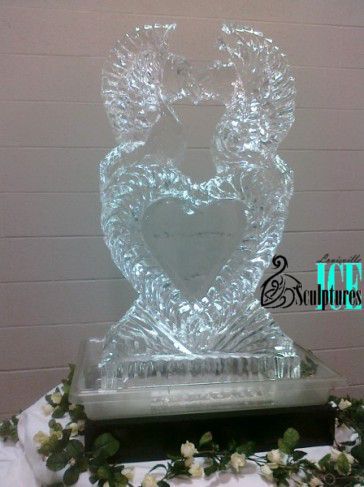 Clear heart-shaped ice sculpture on a pedestal with floral accents and a lit background