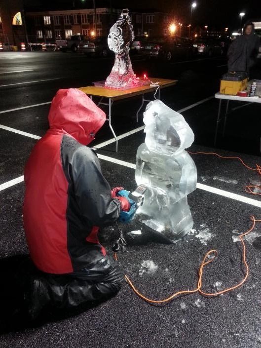 Person in red jacket carving an ice sculpture at night in a parking lot with a glowing worktable