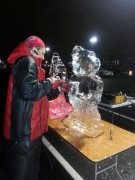 Person in red jacket carving a clear ice bear outdoors at night beside a table