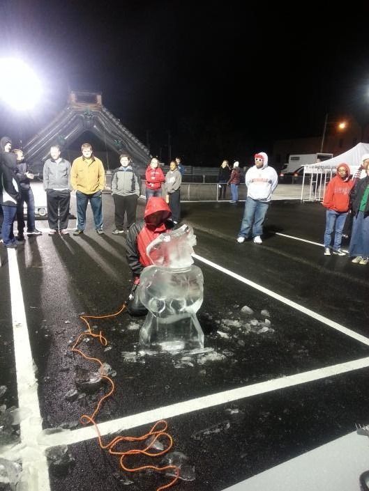 Person carving ice sculpture at night on a wet outdoor court, with spectators nearby and bright lights.