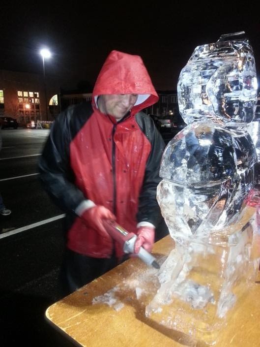 Person in a red hooded jacket carving a clear ice sculpture outdoors at night.