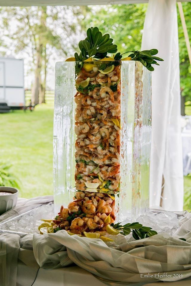 Tall glass vase filled with skewered appetizer bites, garnished with green leaves on a white-draped table outdoors
