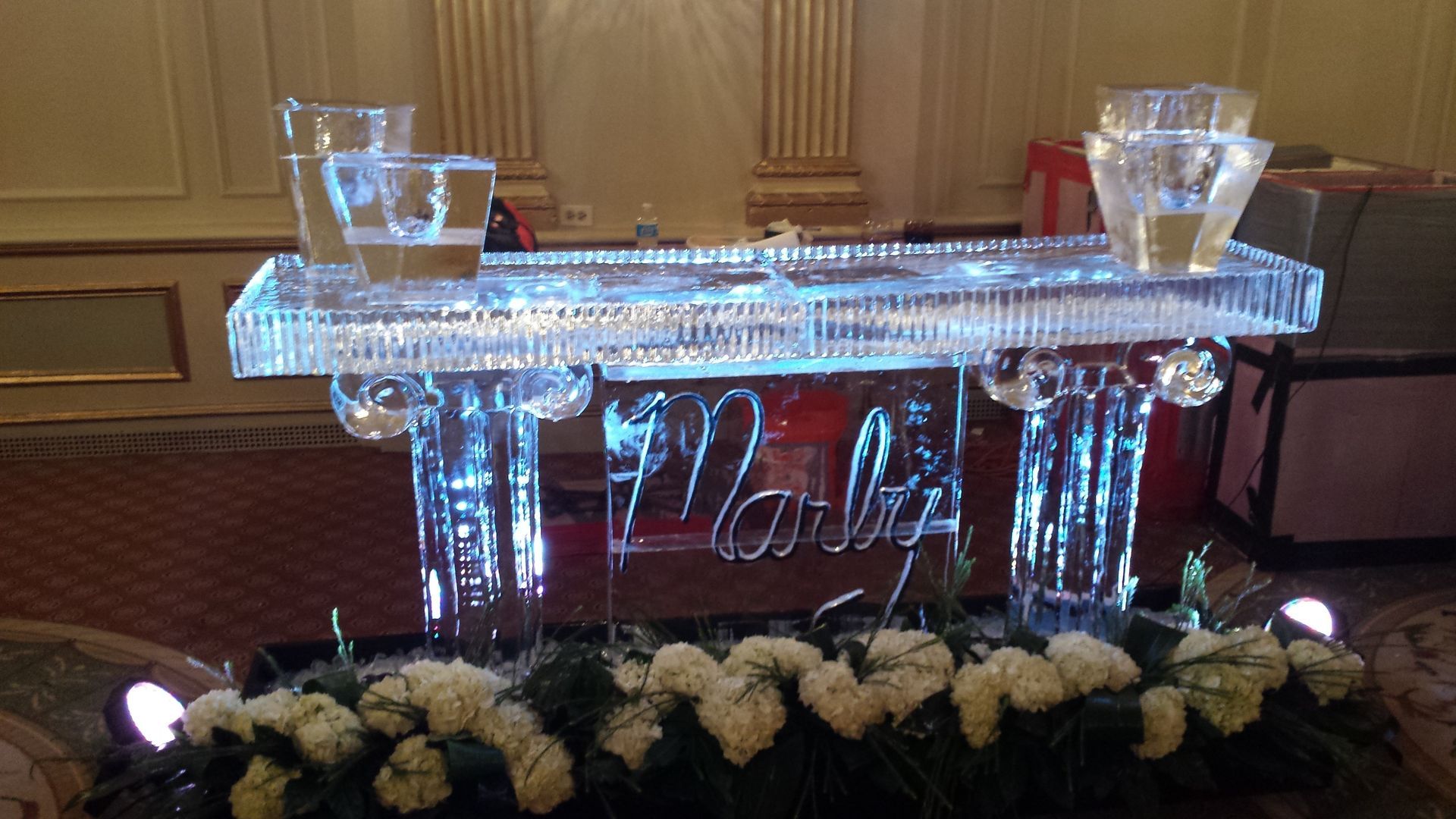 Glowing acrylic bar display with etched name, lit ice buckets, and white floral arrangement at a formal event