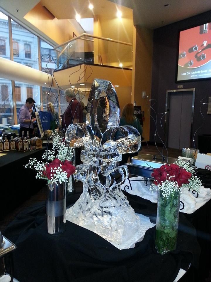 Ice sculpture centerpiece on a banquet table in a warmly lit event hall, with floral vases nearby.