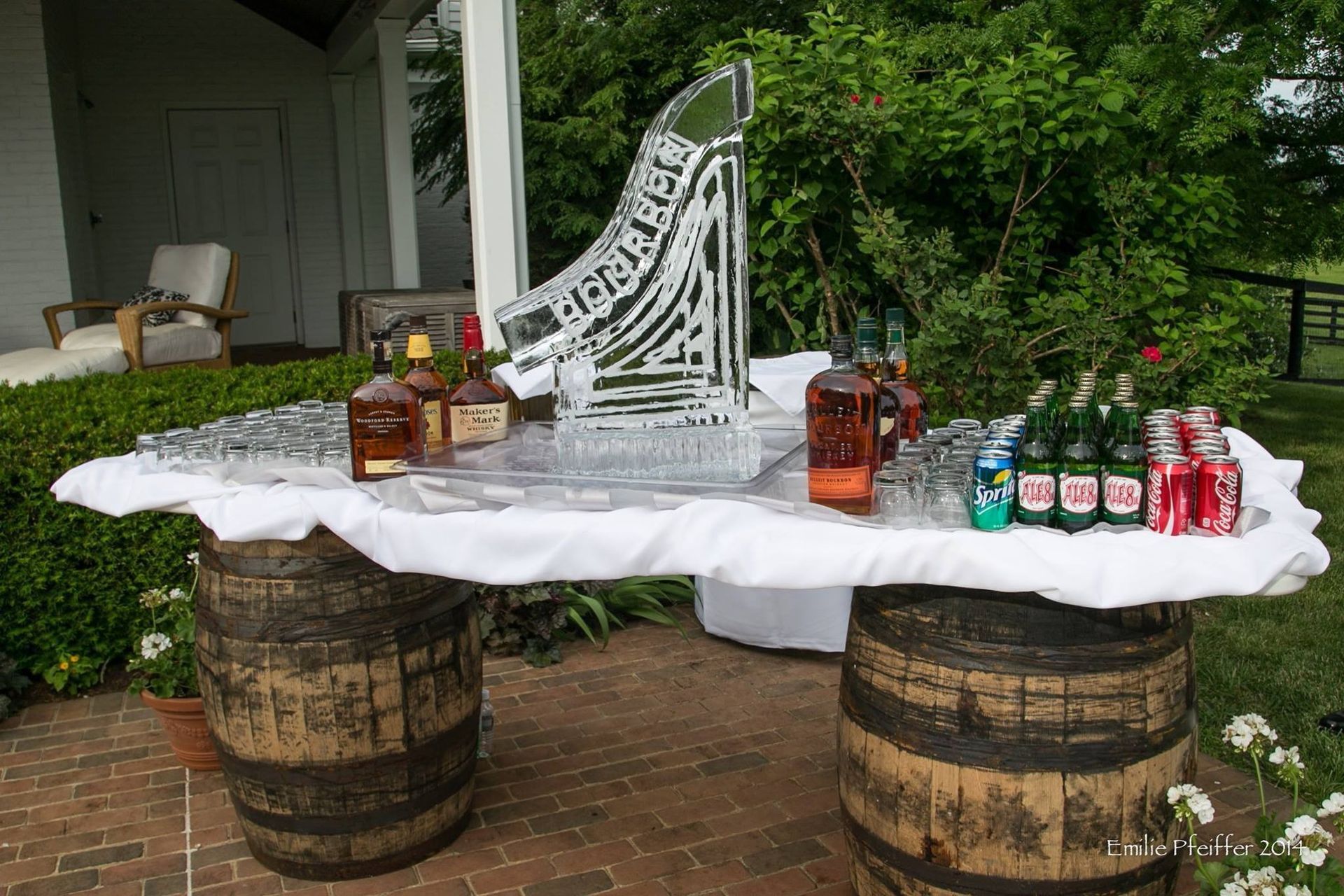 Outdoor refreshment table with white cloth, bottled drinks, and snacks on wooden barrels by a porch