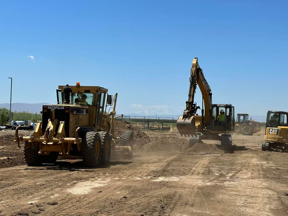 Yellow construction vehicles grading earth on a dirt lot under a clear blue sky.