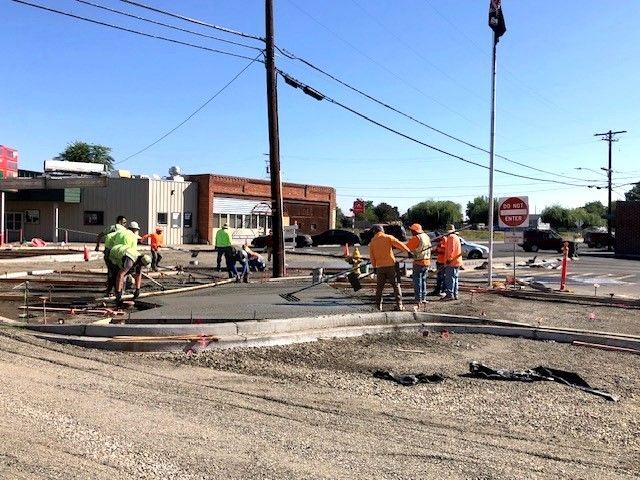 Construction workers pouring concrete on a roadway, with buildings and utility poles in the background.