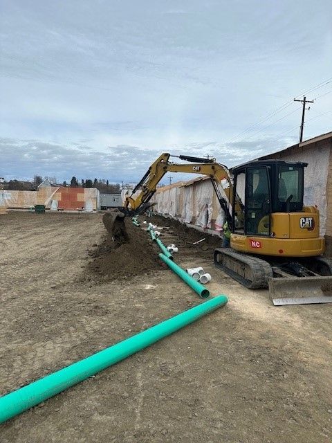 Excavator laying green pipes in a trench next to a building on a cloudy day.