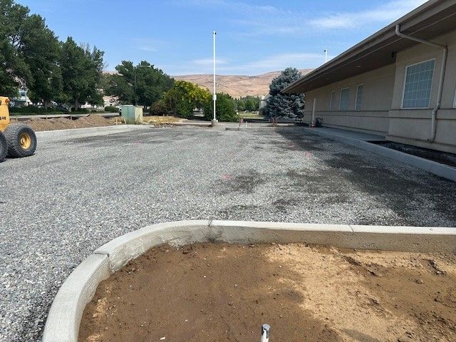 Gravel parking lot next to a building with a newly constructed concrete curb. A flagpole stands in the center.