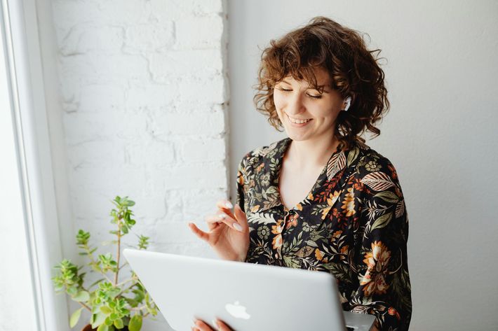 Woman smiling beside a laptop in a bright room, with a leafy plant nearby