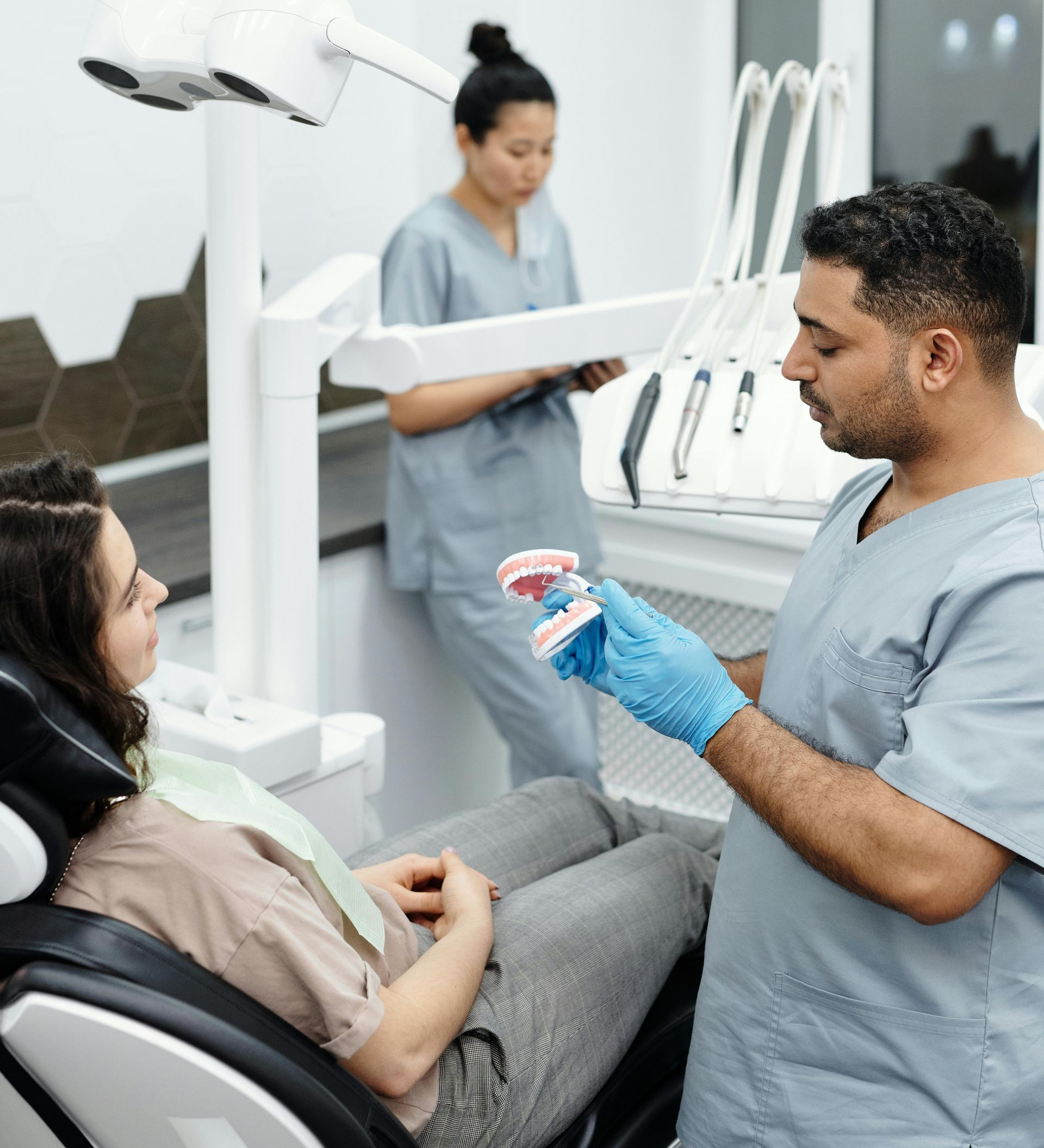 Dentist in blue gloves explaining treatment to patient in a dental chair, assistant in background