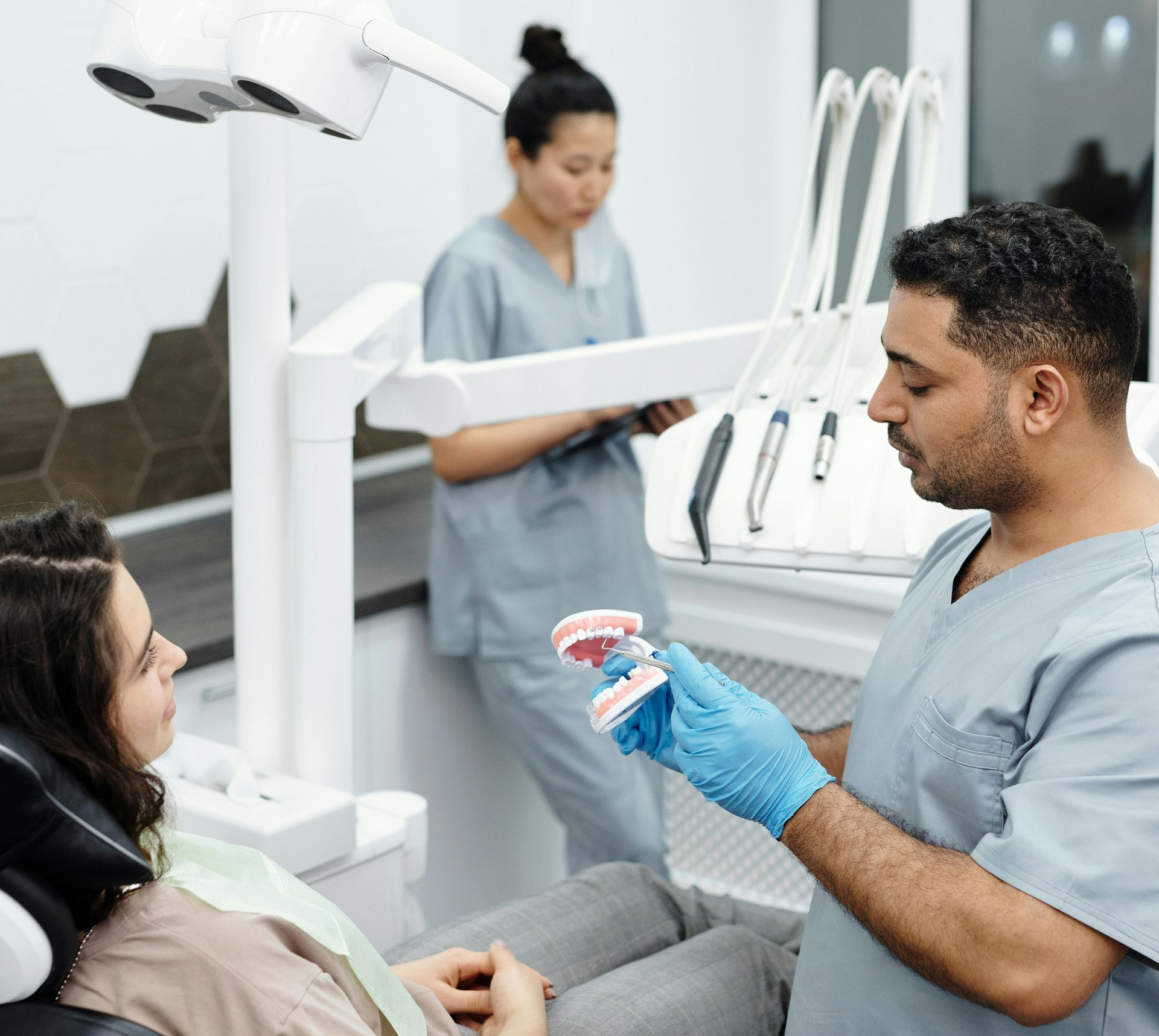 Dentist in blue gloves explaining treatment to patient in a dental chair, assistant in background