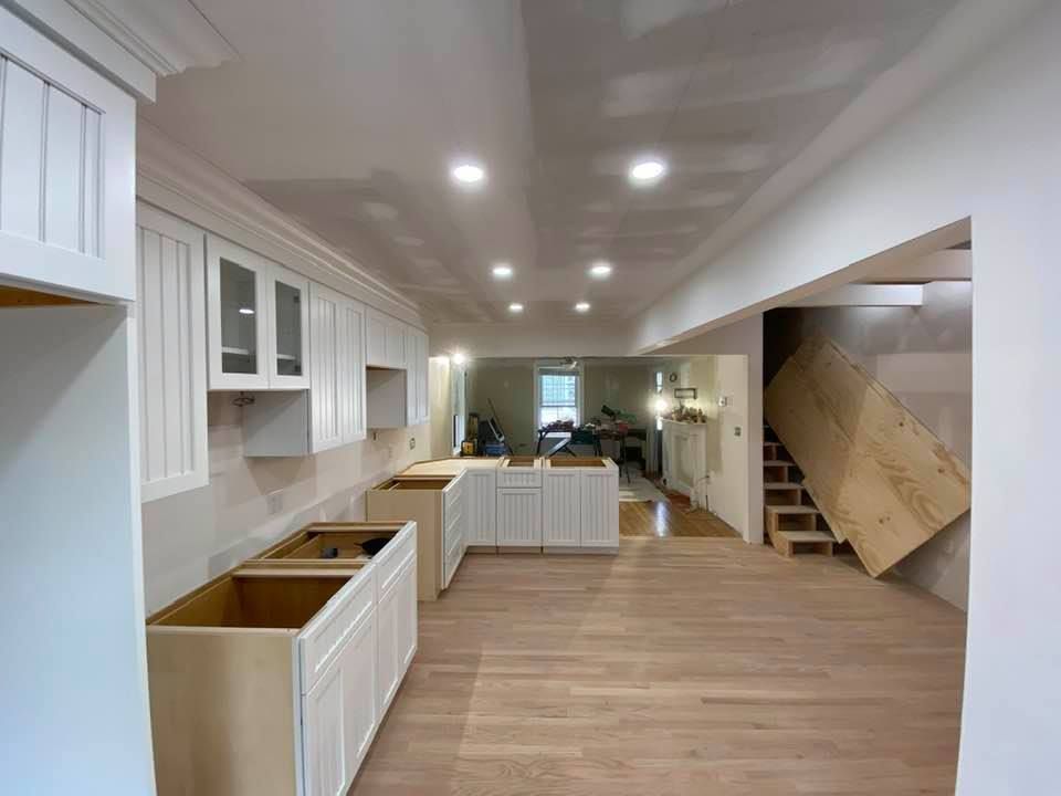 Bright modern kitchen with white cabinets, wood floors, recessed lights, and staircase at the back.