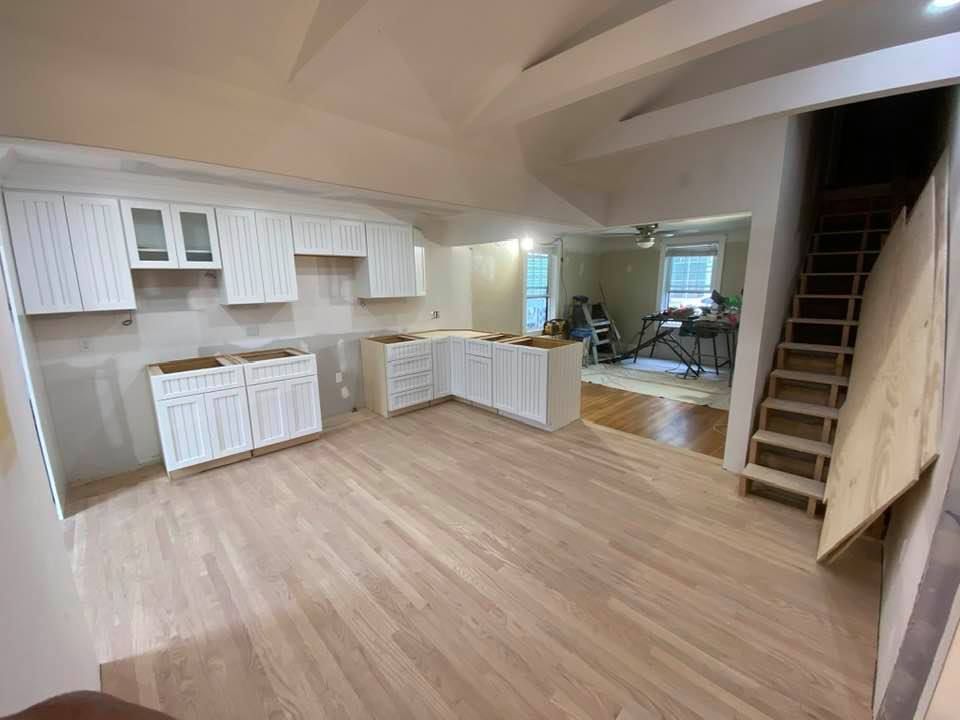 Empty open-plan kitchen and living room with light wood floors, white cabinets, and a staircase on the right