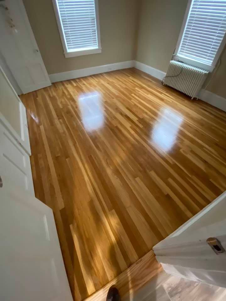 Empty room with glossy hardwood floors and two windows with blinds and a radiator