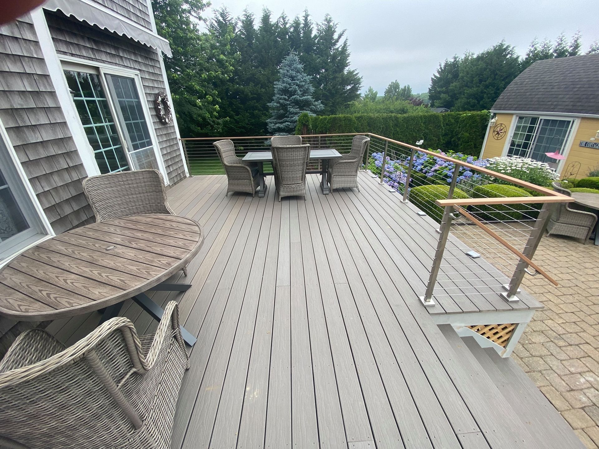 Outdoor deck with wicker seating, dining table, potted flowers, and a view of the yard and trees