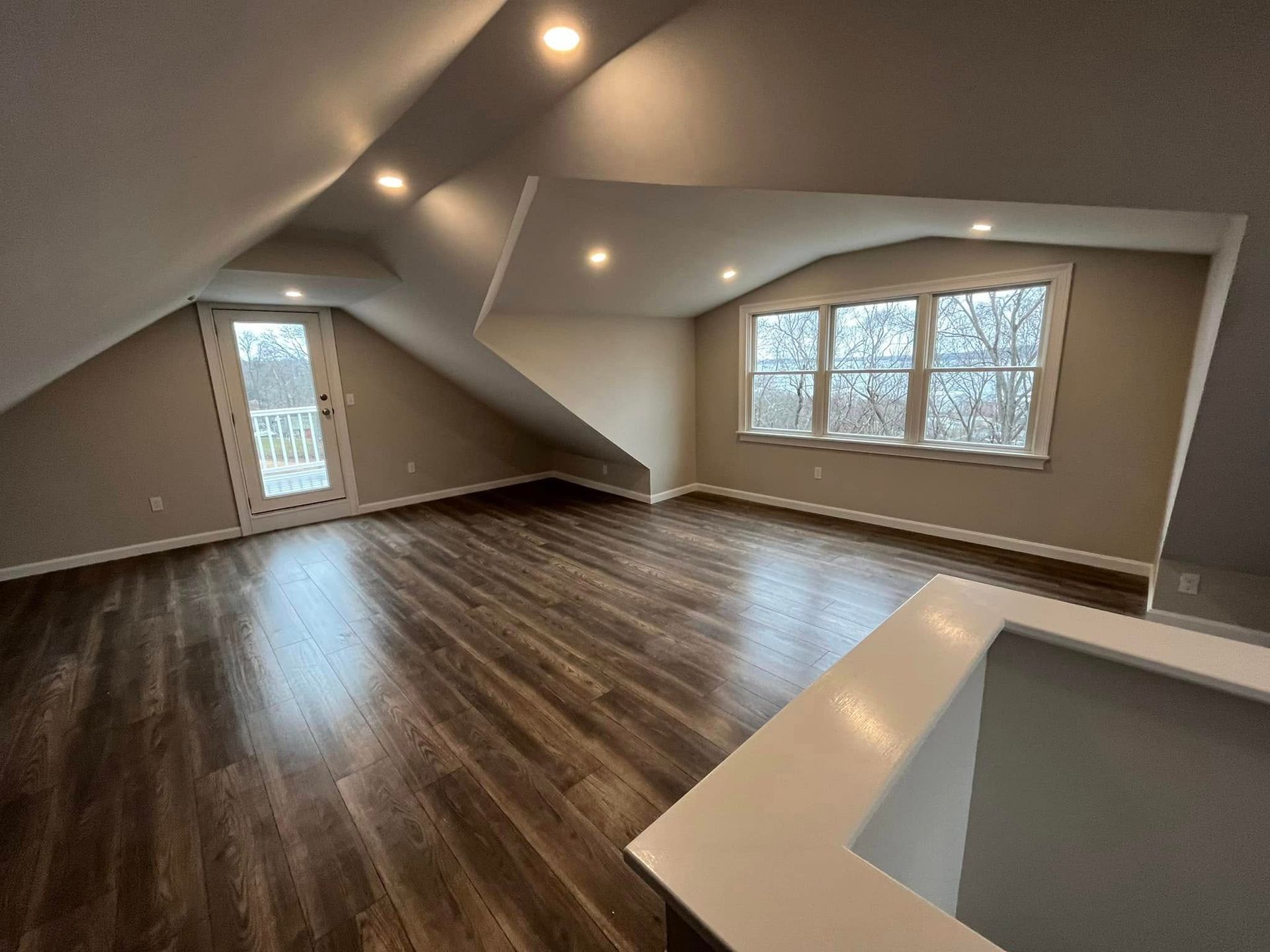 Empty modern loft room with sloped ceiling, dark wood floors, recessed lights, and large windows