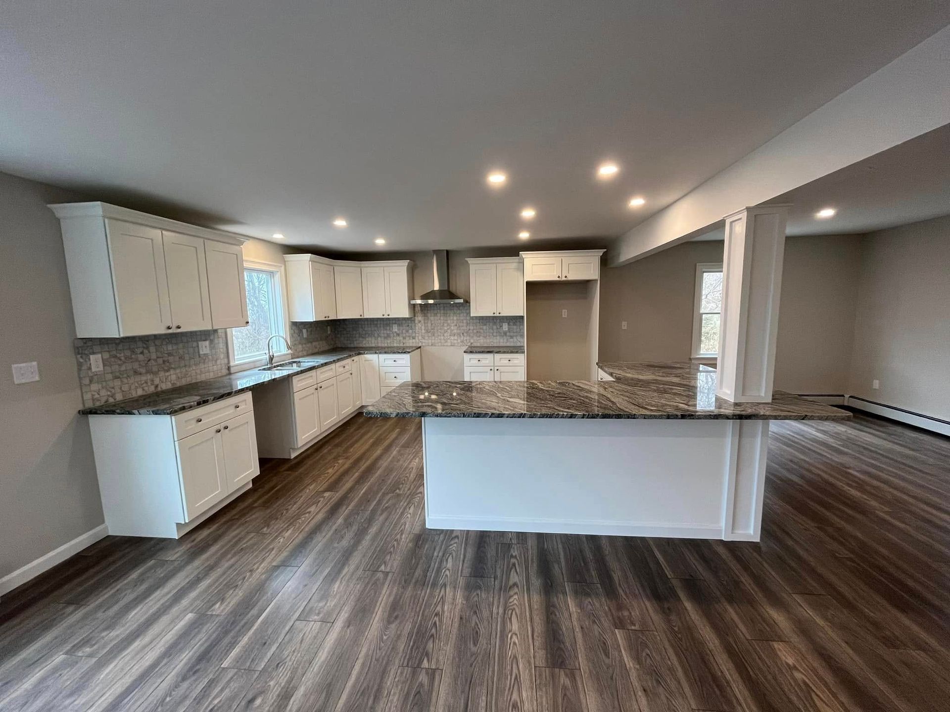 Empty modern kitchen and living area with white cabinets, granite island, and wood-look flooring.