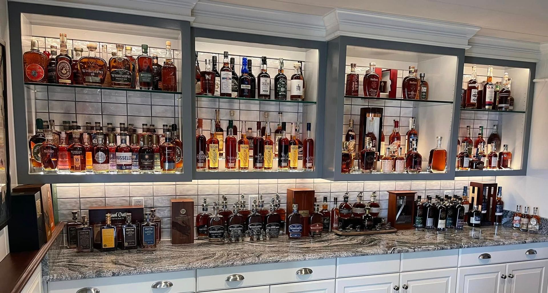 Wall of liquor bottles on illuminated shelves above a bar counter, with glassware and taps below
