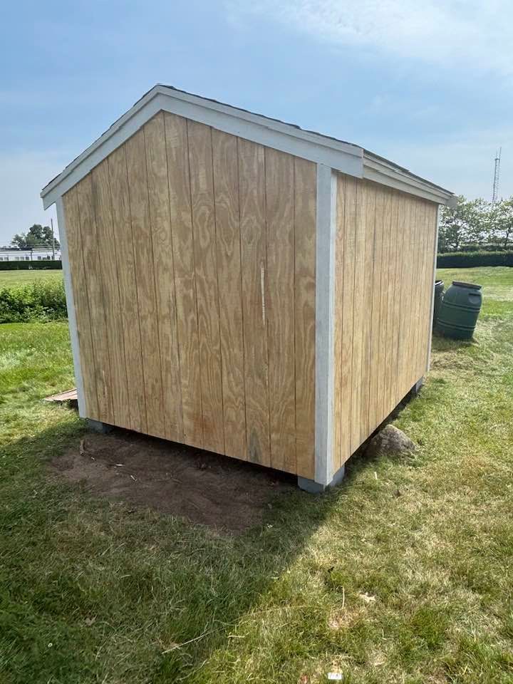 Small unfinished wooden shed with white trim sitting on a grassy field under a clear sky