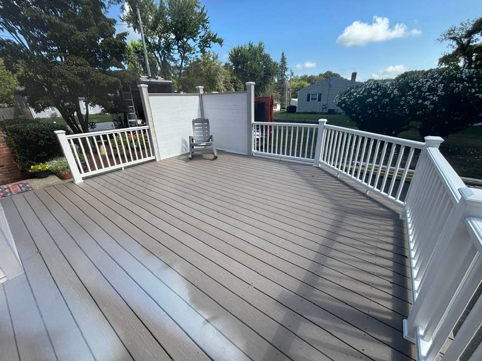 Spacious gray composite deck with white railings and chairs, overlooking a backyard under a blue sky.