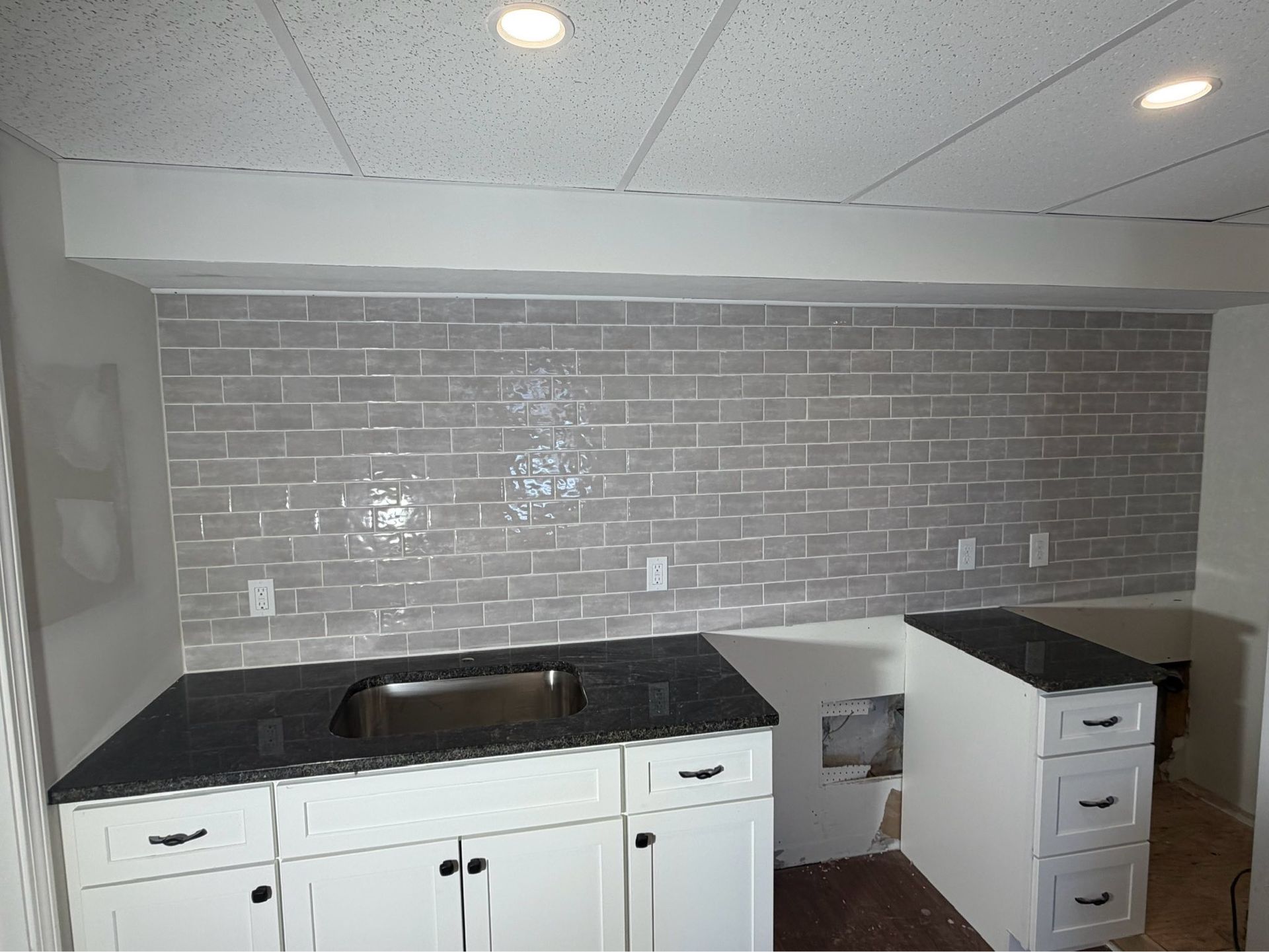 Kitchen with white cabinets, black countertop, brick-pattern backsplash, and sink under bright ceiling lights