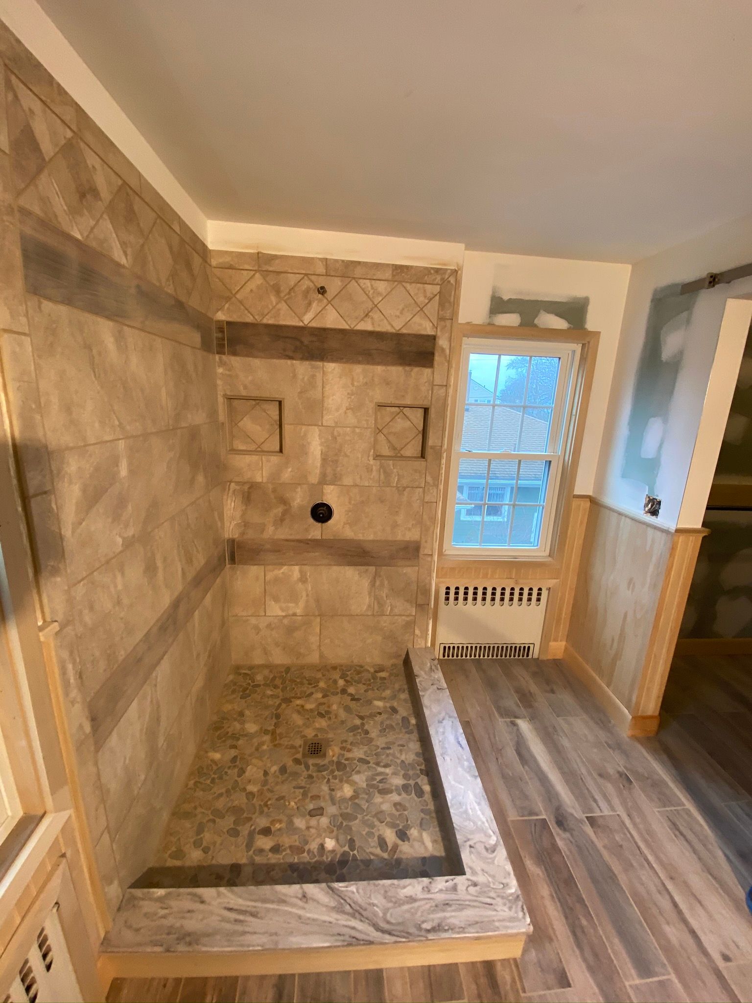 Small tiled shower stall with a bench, beige walls, and a window beside a radiator.