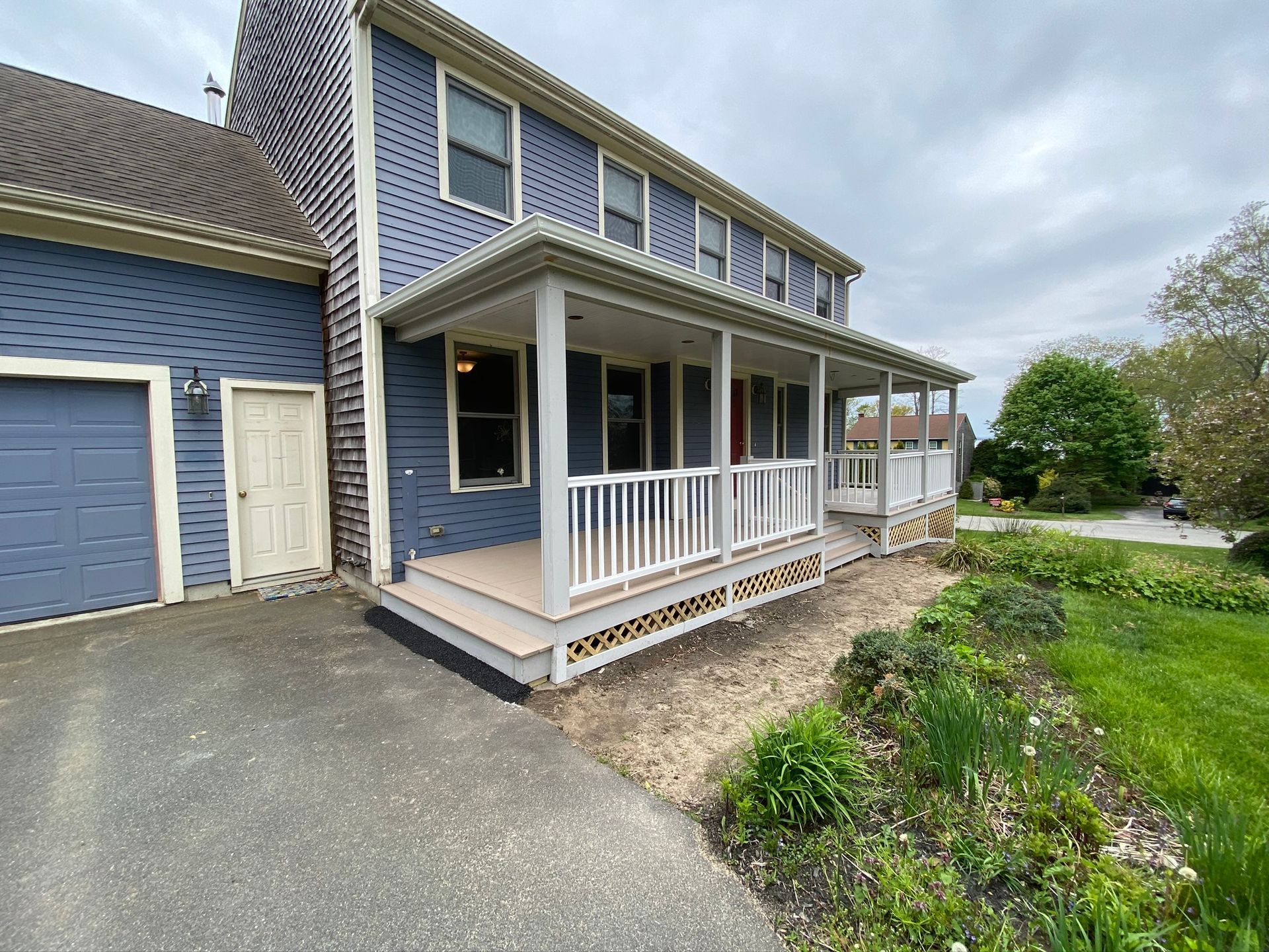 Blue house with white porch railing, garage, and driveway beside a grassy yard under cloudy sky