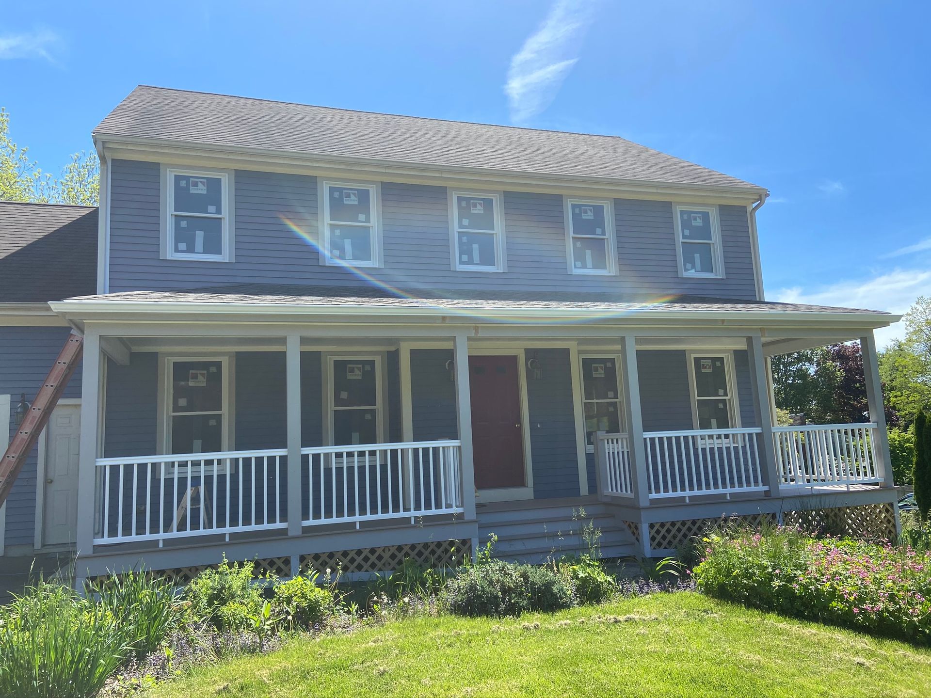 Two-story gray house with white porch railings and flower beds under a bright blue sky