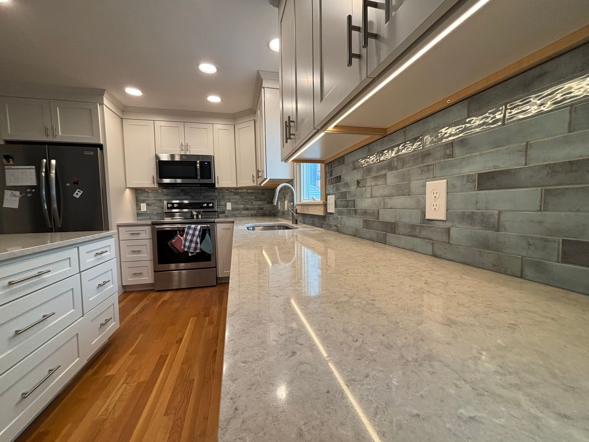 Bright modern kitchen with white cabinets, stainless appliances, wood floor, and long speckled countertop.