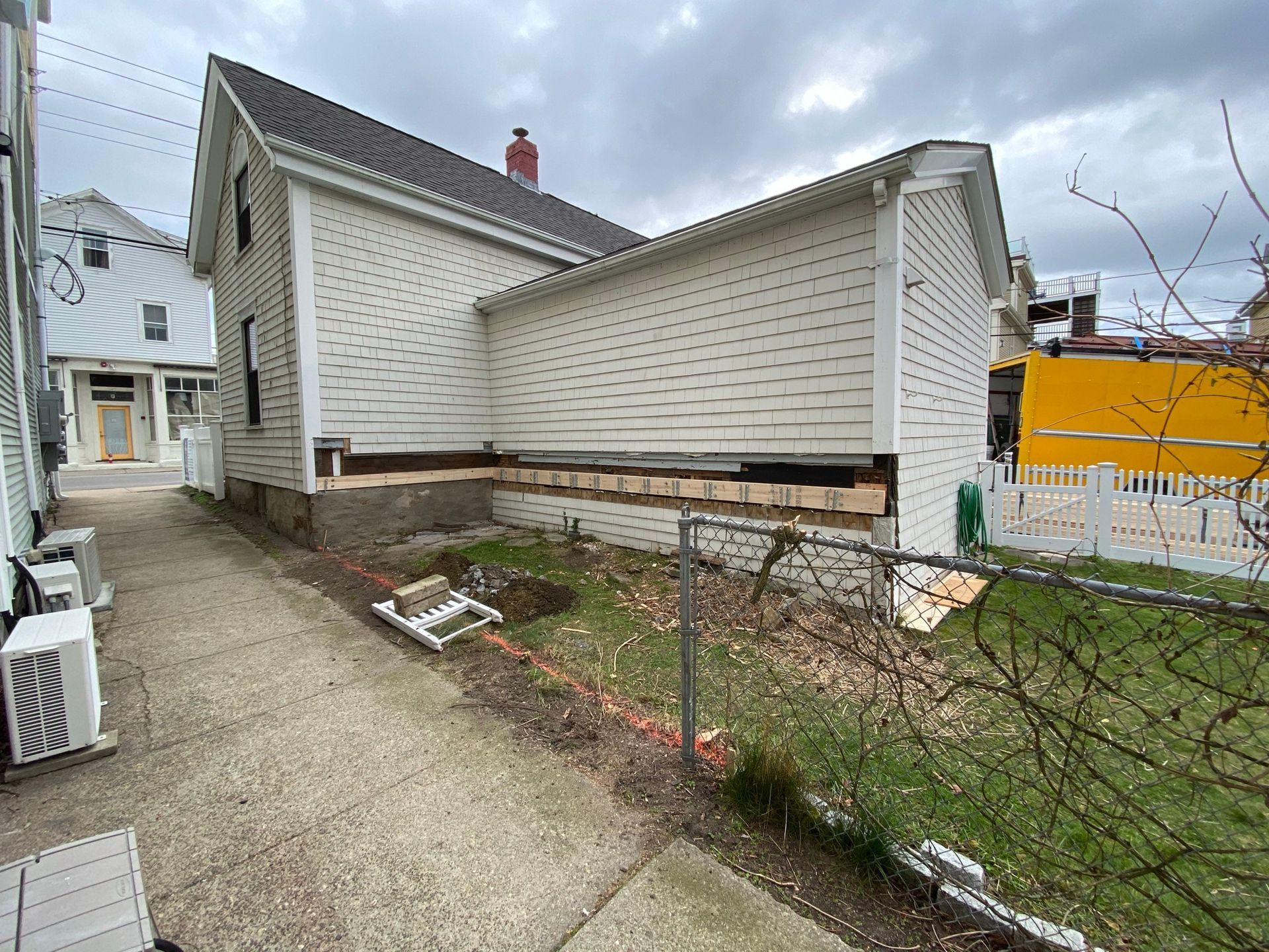 Side view of a weathered white house with a gravel driveway, small yard, and a yellow truck in the background