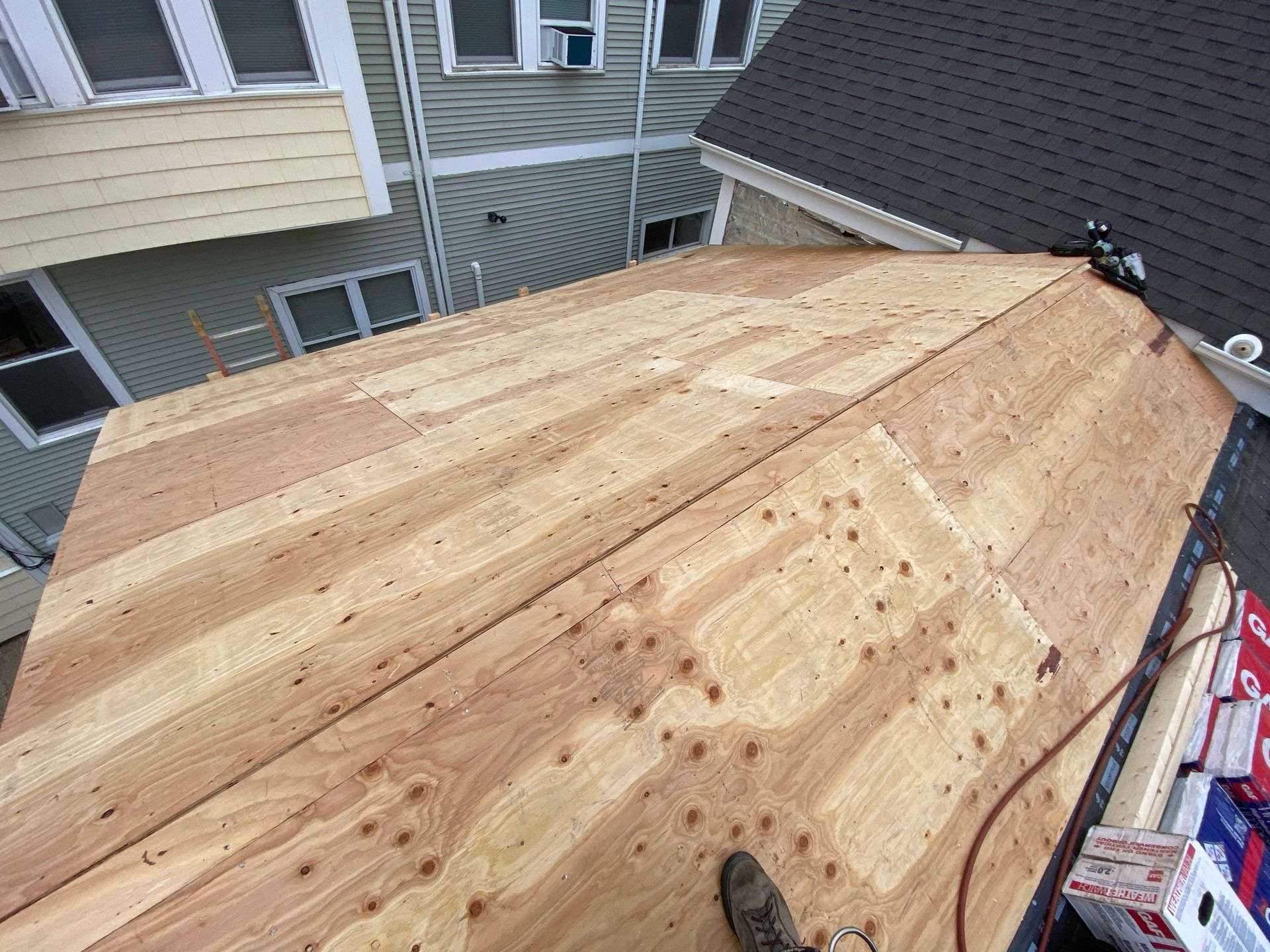 Roof being reroofed with plywood sheathing on a house, viewed from above, with nearby homes visible.