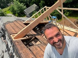 Man smiling beside a partially rebuilt wooden roof frame on a house under construction
