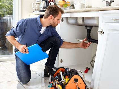 Plumber crouches under a kitchen sink, inspecting pipes with a blue folder and tools nearby.